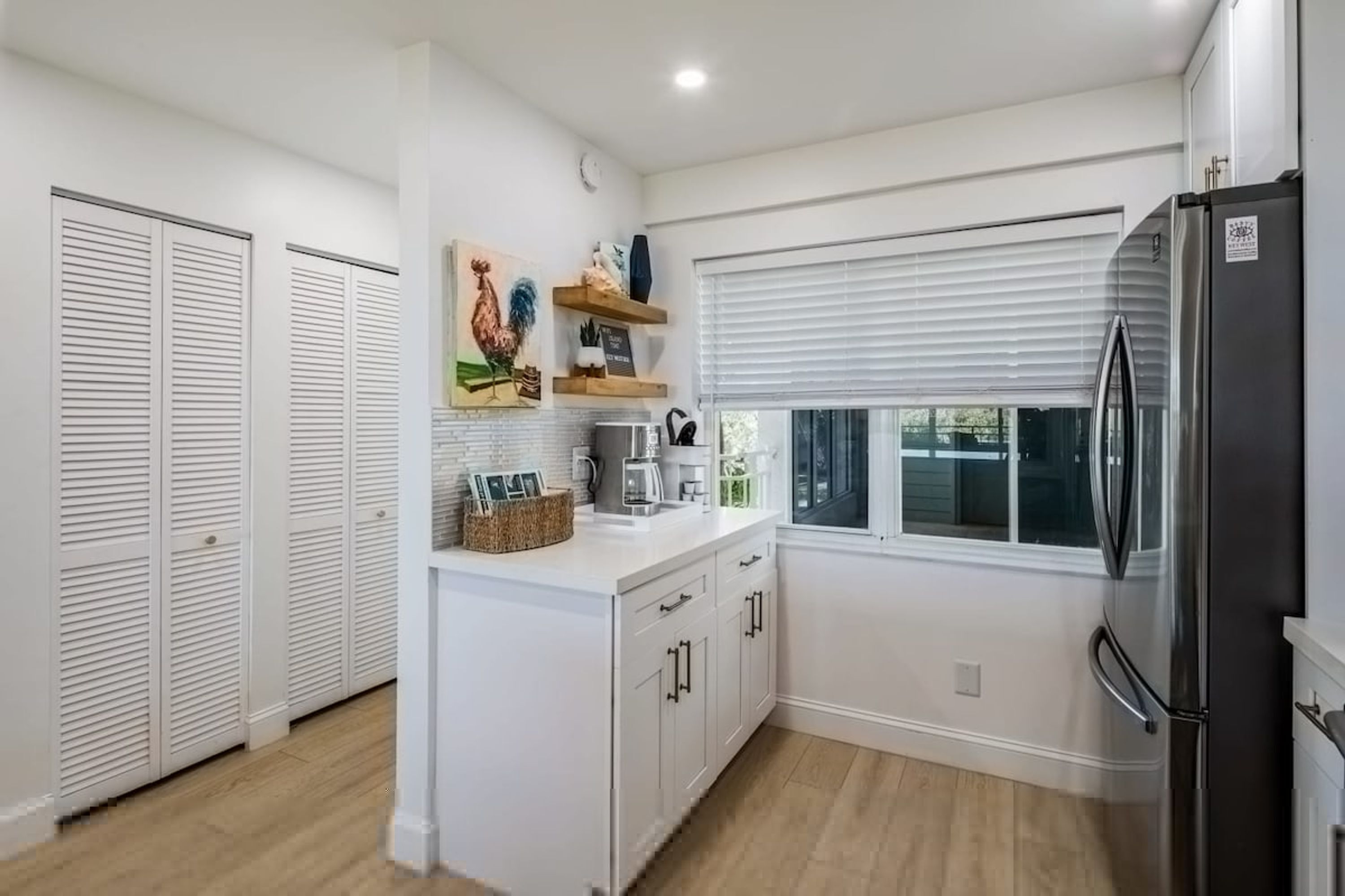 A modern kitchen with white cabinetry, a stainless steel refrigerator, and a window with blinds.