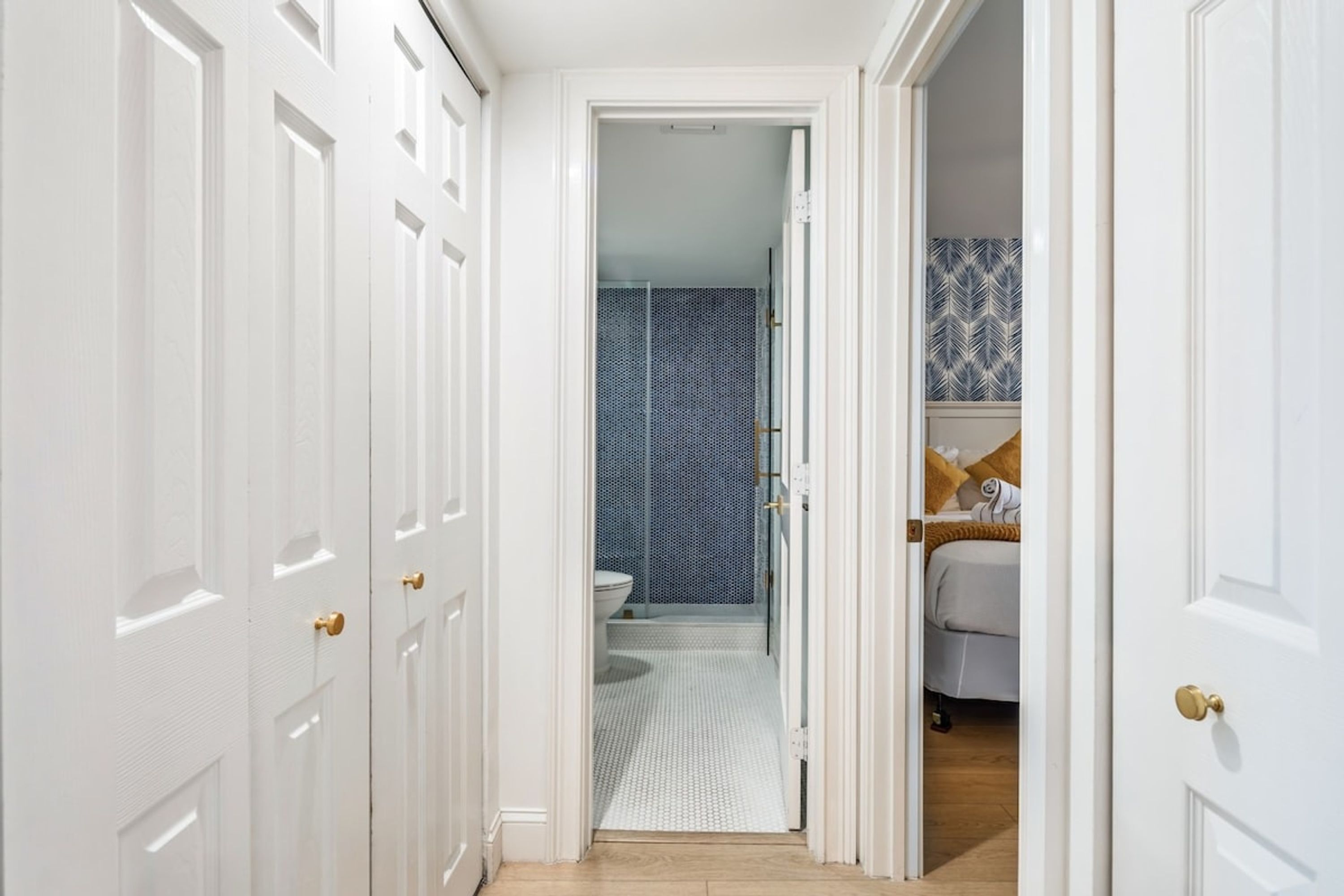 A hallway leading to a bathroom with a shower and toilet, and a bedroom with a bed and a patterned wall.
