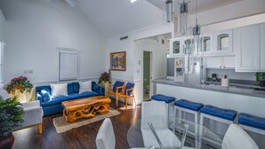 Ein modernes Wohnzimmer mit einem blauen Sofa, einem Holztisch und einem Glastisch. Im Hintergrund ist eine Küche mit weißen Schränken und einer Bar erkennbar.