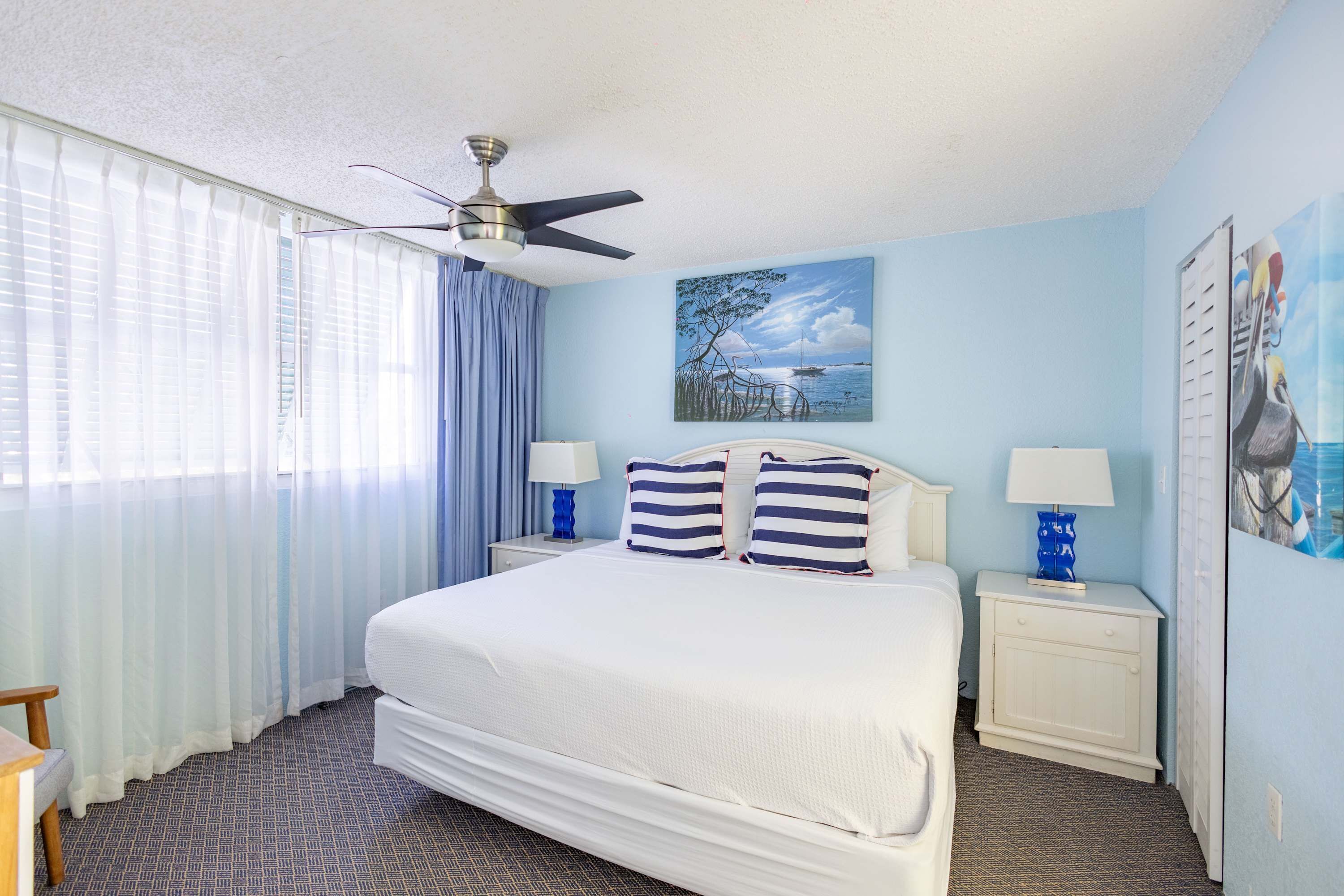 A hotel room with a large bed, two bedside tables with lamps, a ceiling fan, and a painting of a coastal scene.