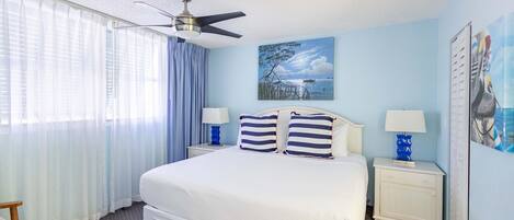 A hotel room with a large bed, two bedside tables with lamps, a ceiling fan, and a painting of a coastal scene.