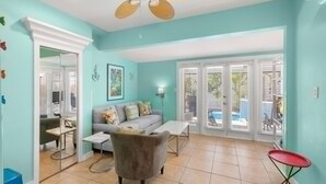 A living room with a sofa, coffee table, and a red chair. There is a ceiling fan, a large window with blinds, and a door leading to another room. - Poolside Paradise by Brightwild Heated Pool (Key West)