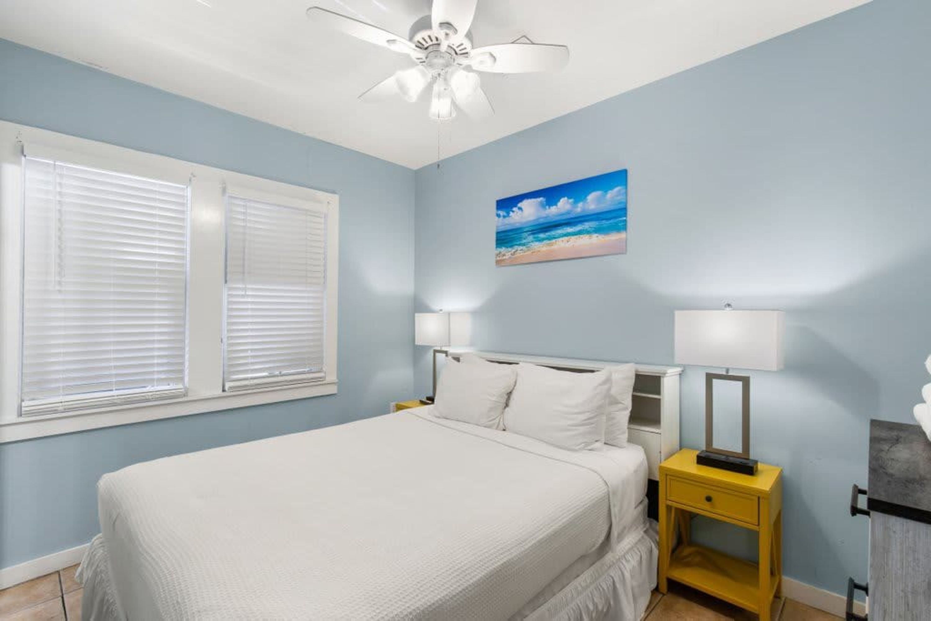 Une chambre à coucher avec un lit, deux lampes de chevet, une table de chevet, un ventilateur de plafond et un tableau sur le thème de la plage accroché au mur.