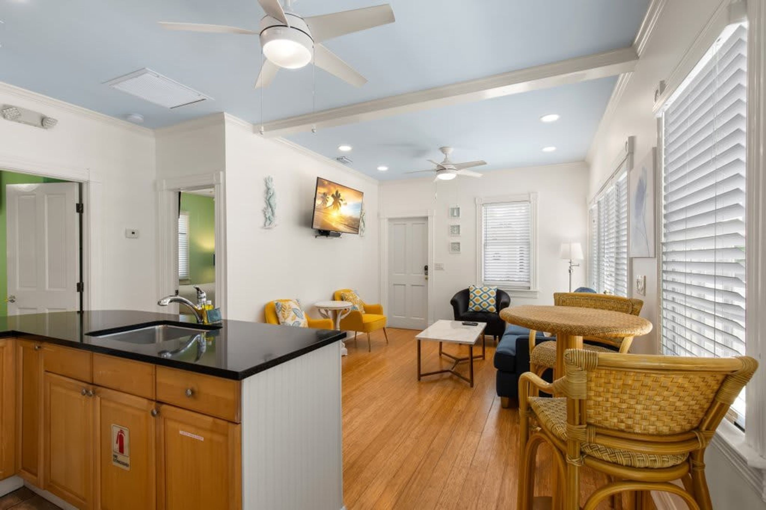A modern kitchen with a black countertop and wooden cabinets, a dining area with wicker chairs and a small table, and a ceiling fan.