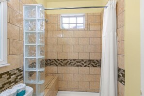 A bathroom with beige tiles, a glass shower enclosure, a window, and a white shower curtain.