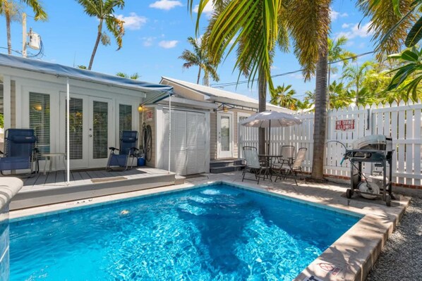 A swimming pool with lounge chairs and umbrellas, surrounded by a white fence and palm trees.