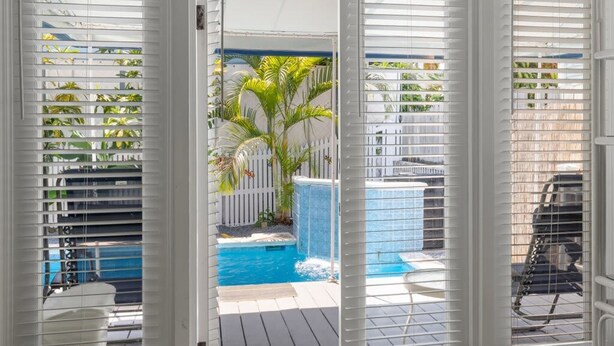 Vista a través de persianas blancas de un área de piscina con jacuzzi, rodeada de palmeras y una valla blanca.