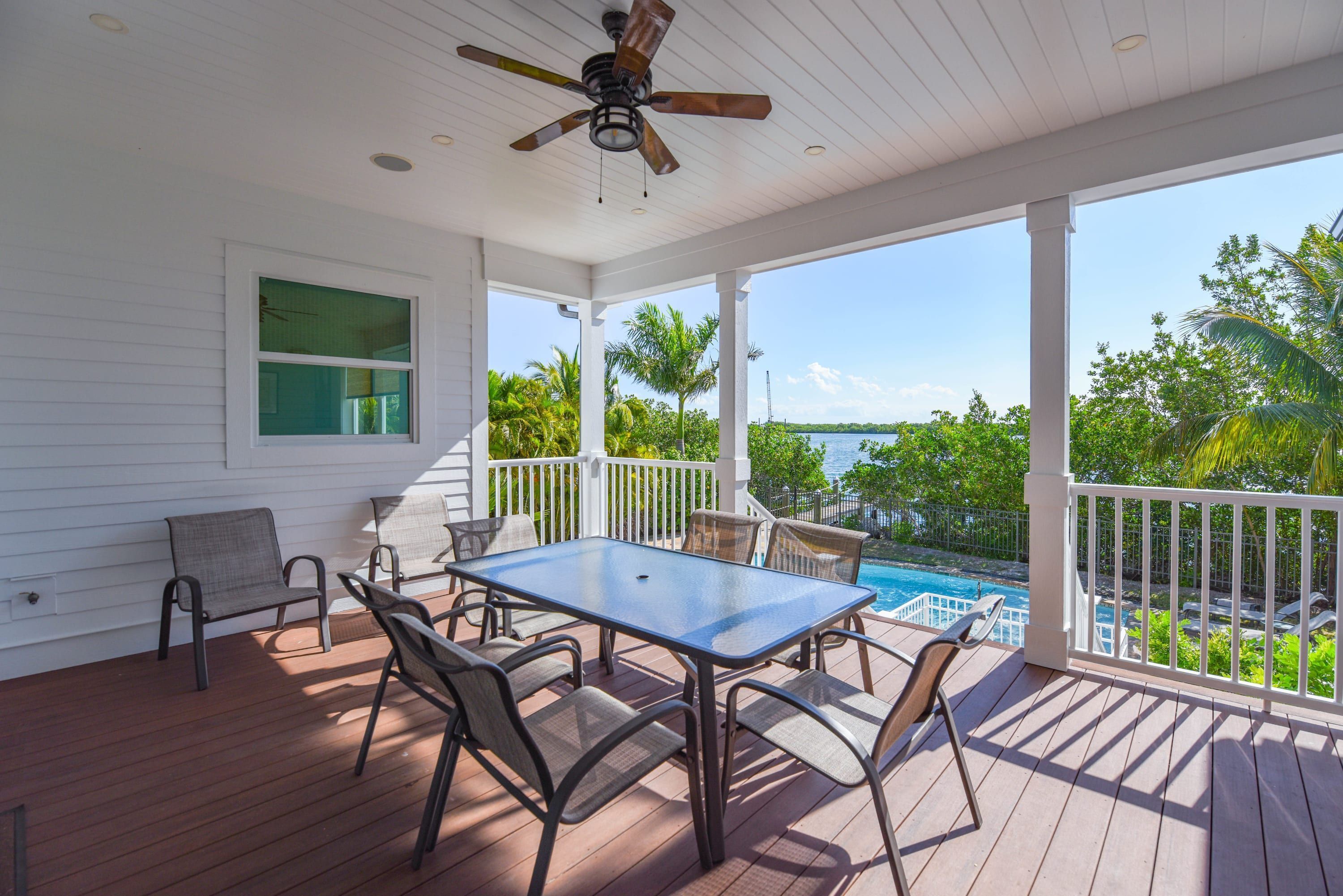 A covered outdoor area with a table and chairs, a ceiling fan, and a view of a swimming pool and greenery.