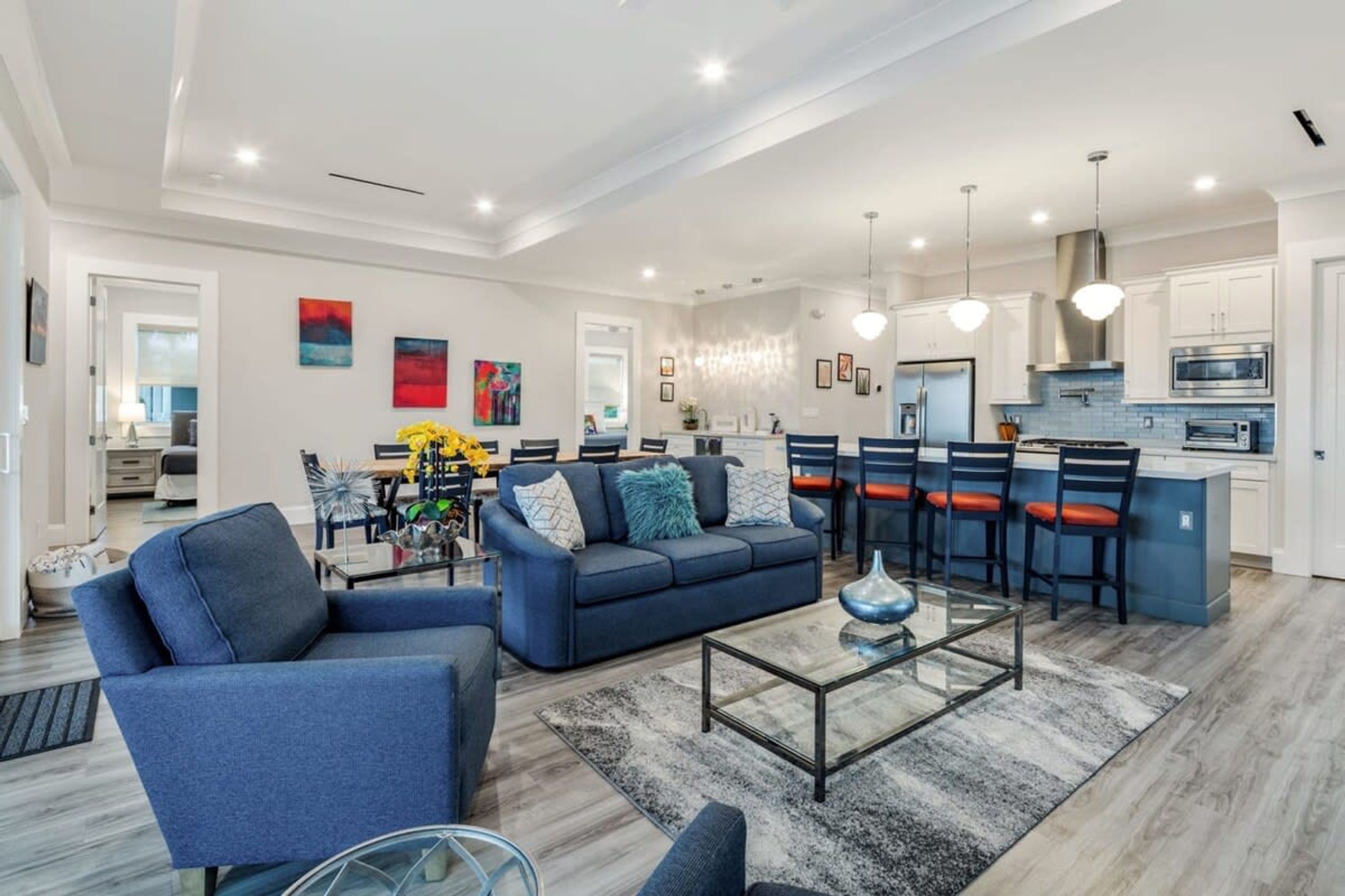 A modern living room with a blue sofa, a glass coffee table, and a dining area with a blue island and red chairs.