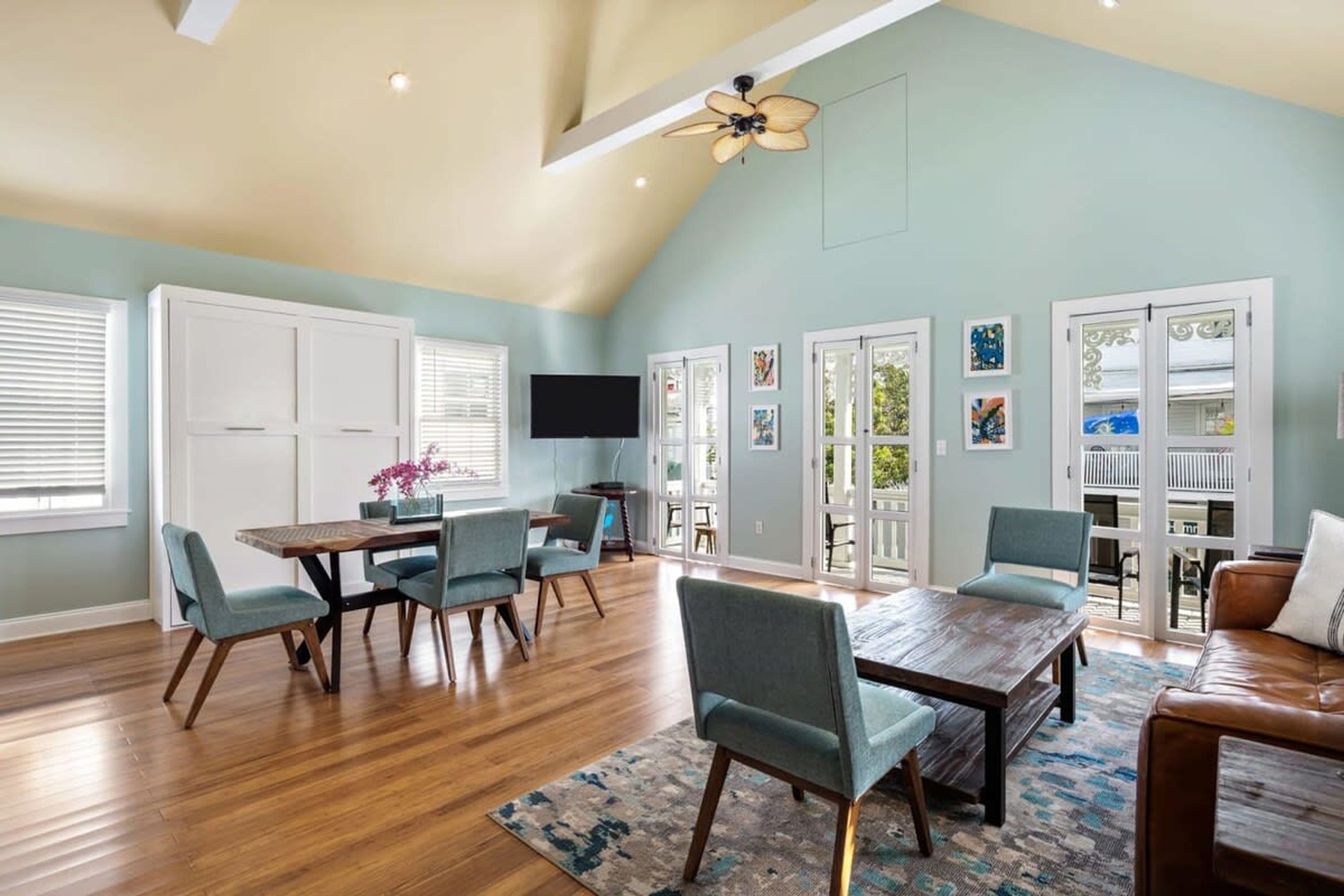 A spacious living room with a high ceiling, wooden flooring, and a large dining table with chairs. There are sliding glass doors leading to a balcony.