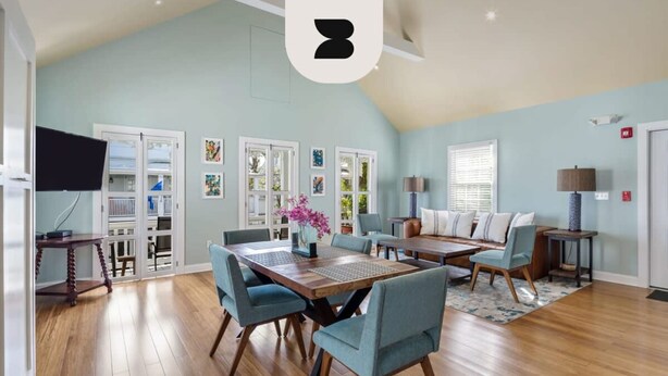 Een ruime woonkamer met een hoge plafond, een eethoek met een houten tafel en blauwe stoelen, en een gezellige zithoek met een bank en fauteuils.