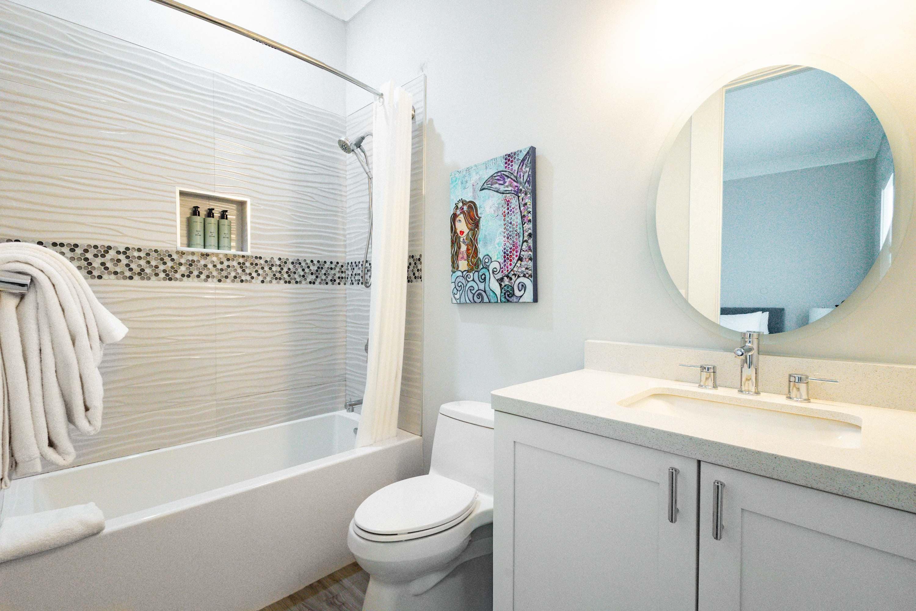 A bathroom with a bathtub, toilet, and a sink with a mirror. There is a towel rack with towels and a shelf with toiletries.