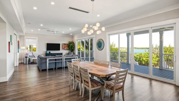 A spacious living area with a wooden dining table, comfortable seating, a flat-screen TV, and large windows offering a view of the outdoors. - Spindrift House by Brightwild Huge Pool Dock (Key West)