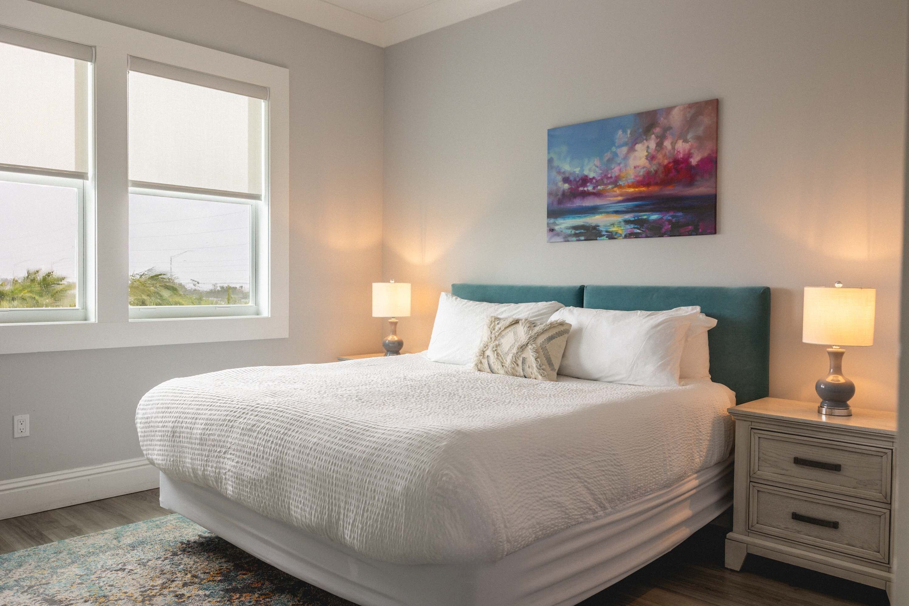 A bedroom with a large bed, two bedside tables with lamps, a window, and a painting on the wall.