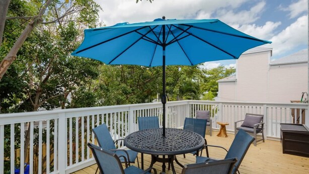 Eine Terrasse mit einem runden Tisch und Stühlen, einem blauen Sonnenschirm und einem weißen Geländer.
