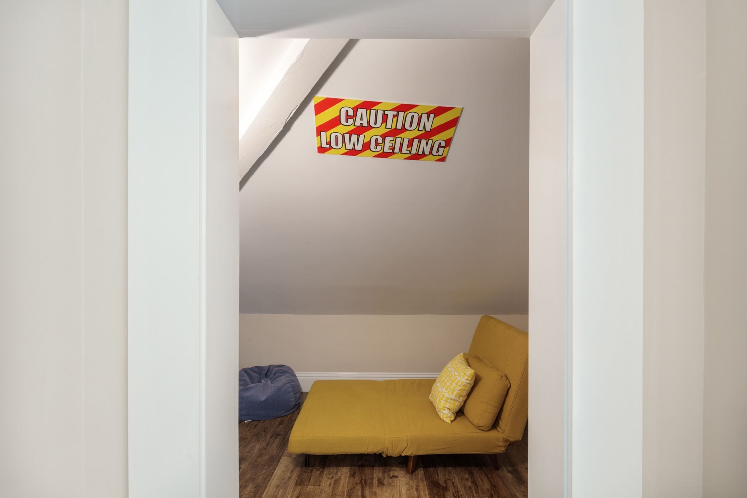 A room with a low ceiling warning sign, a mustard-colored sofa, and a blue bean bag chair.