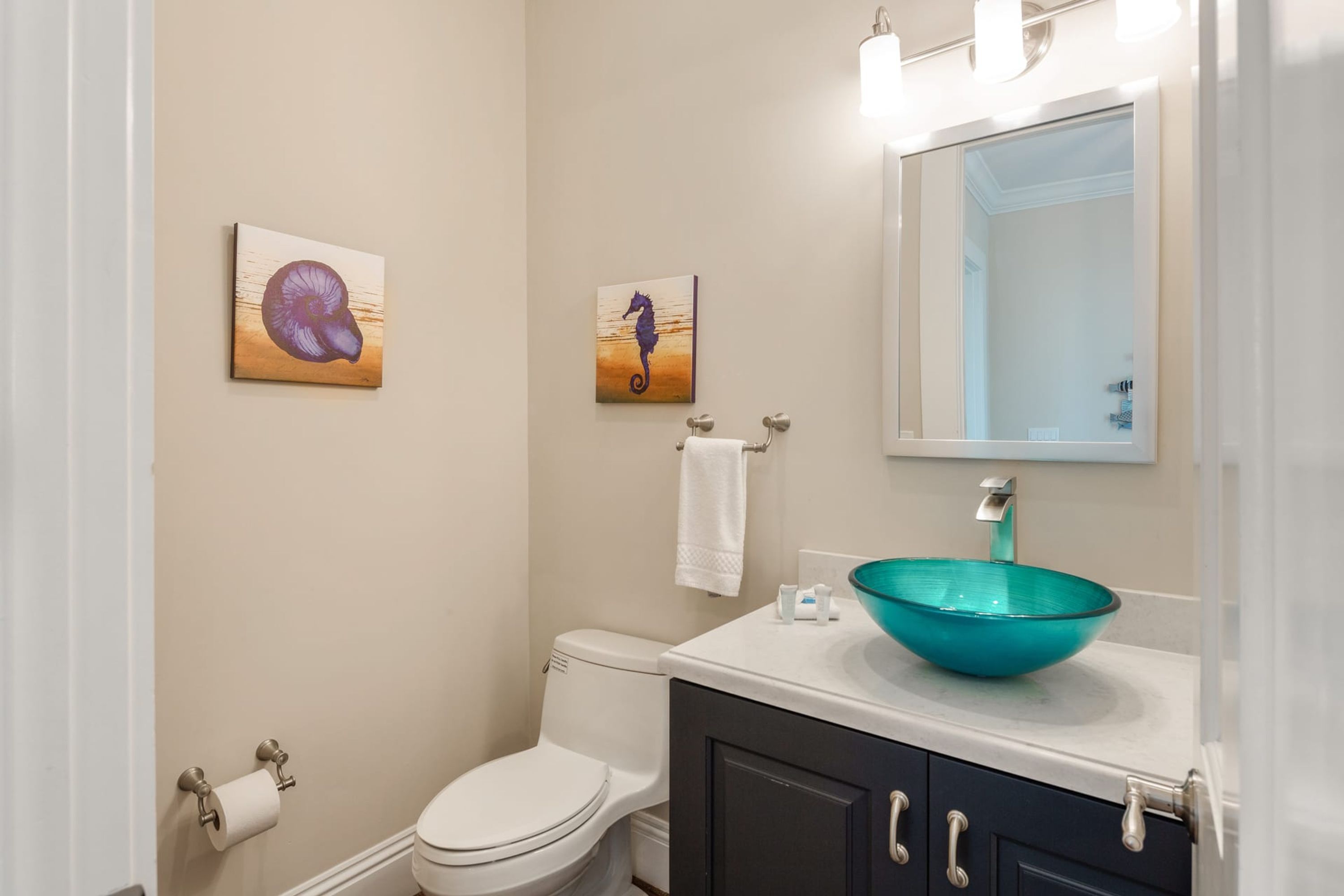 Une salle de bain comprenant des toilettes, un lavabo avec un récipient en verre bleu et deux tableaux accrochés au mur.