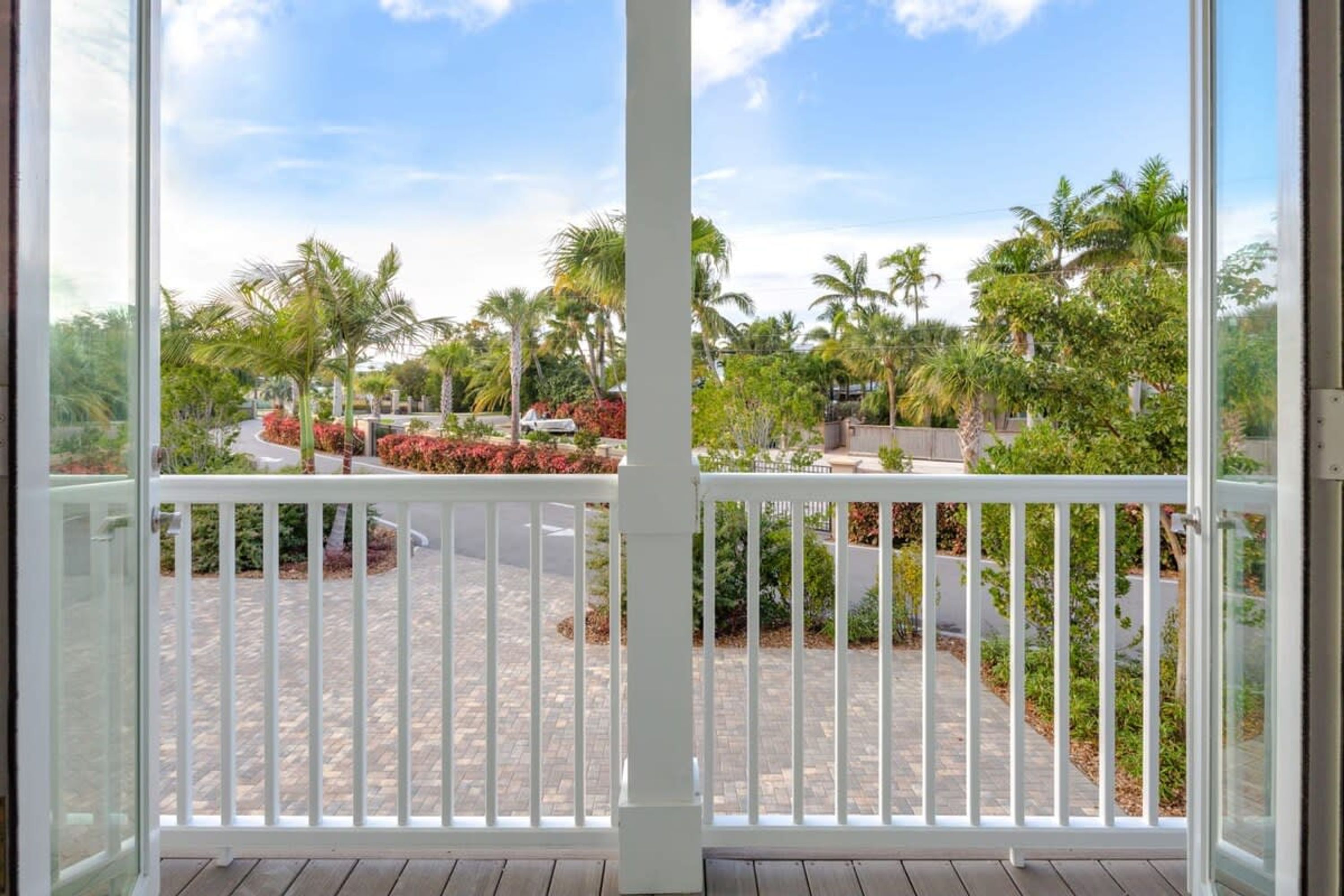 A view from a balcony with a white railing, overlooking a paved area with palm trees and red flowers.