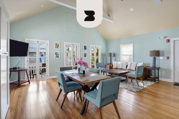 Een ruime woonkamer met een hoge plafond, houten vloer, een eettafel met stoelen, een bank en een televisie.