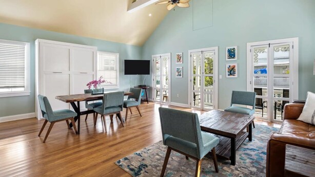 A spacious living room with a high ceiling, wooden flooring, and a large dining table with chairs. There are sliding glass doors leading to a balcony. - Sky Suites Retreat by Brightwild 2 Stunning Condos (Key West)