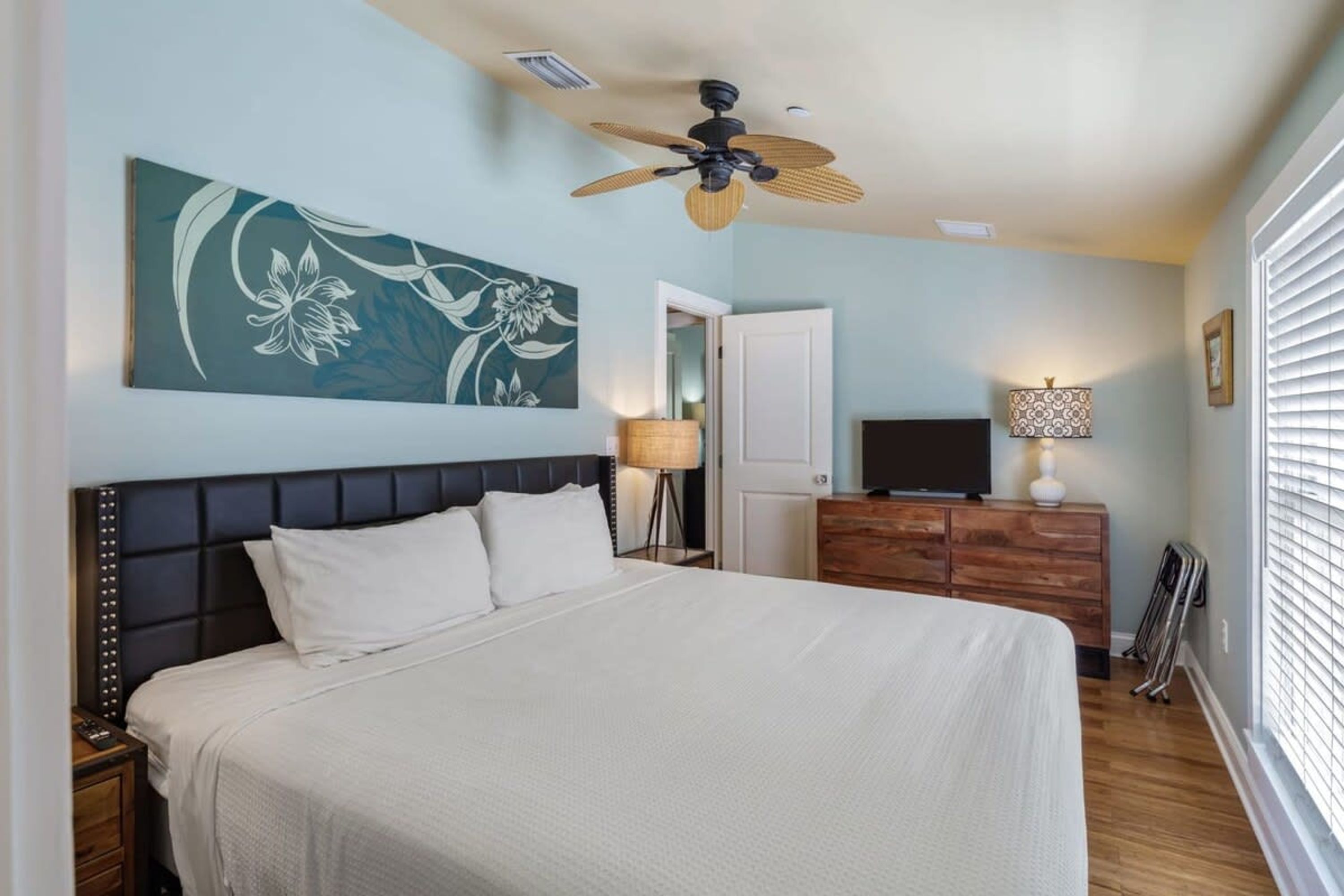 A bedroom with a bed, a ceiling fan, a TV, a lamp, and a painting on the wall.
