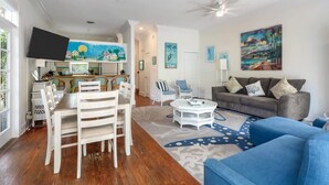 A spacious living area with a dining table and chairs, a sofa, and a coffee table. There are ceiling fans, artwork on the walls, and a TV mounted on the wall. - Sunnyside Up by Brightwild Pool Parking (Key West)