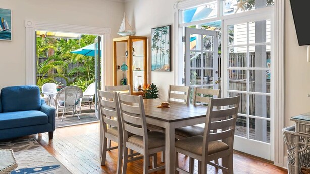 A dining area with a wooden table and chairs, a blue sofa, and a view of the outdoors through a glass door. - Sunnyside Up by Brightwild Pool Parking (Key West)