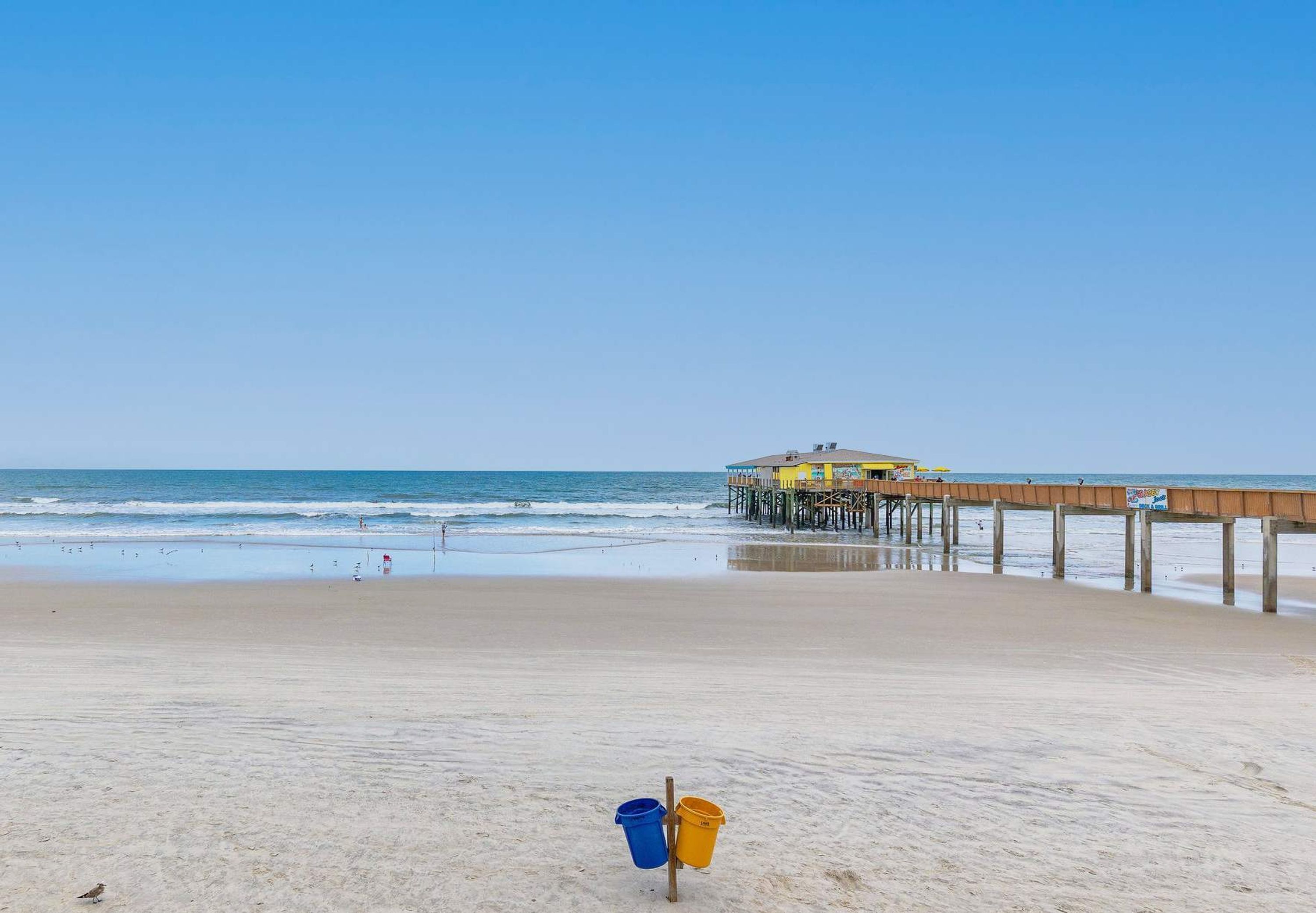 A beach with a wooden pier extending into the ocean, a clear sky, and two buckets on the sand.