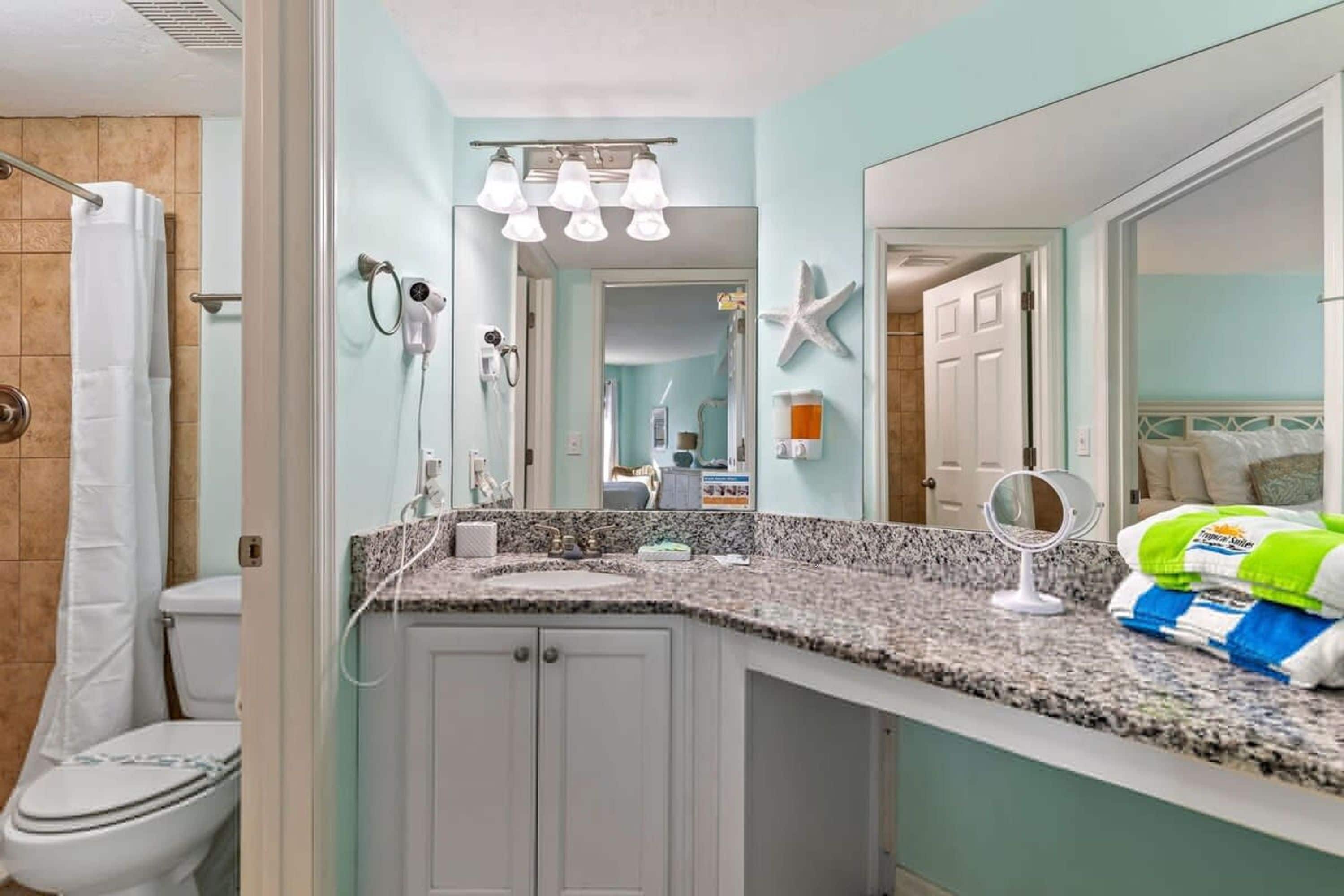 Une salle de bain avec des toilettes blanches, un espace douche, un comptoir en granit, un lavabo, un miroir et un porte-serviettes.