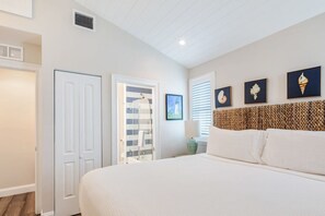 A bedroom with a large bed, a door, a window with blinds, and framed artwork on the wall. - Southernmost View by Brightwild Pool H20 Views (Key West)