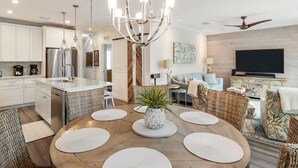 A modern kitchen with stainless steel appliances, a dining area with a round table and placemats, wicker furniture, and a large TV in the living room. - Southernmost View by Brightwild Pool H20 Views (Key West)
