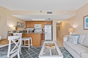 A compact living space with a kitchenette, a dining area with white chairs, a sofa with a patterned cushion, and a coffee table. - Top Floor Oceanfront Condo w Pool Beach Access (Daytona Beach Shores)