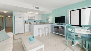A modern kitchen with white appliances, a dining area with teal chairs, and a TV on a stand. - Updated Ocean Front Condo Aquamarine Suite 201 (Daytona Beach Shores)