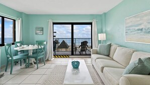 A living room with teal walls, a white sofa, a dining table with chairs, and a view of the beach through a glass door. - Updated Ocean Front Condo Aquamarine Suite 201 (Daytona Beach Shores)