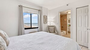 Ein Schlafzimmer mit einem großen Bett, einem Fenster mit Blick auf den Strand, einem Sessel und einem durchgehenden Türrahmen einsehbaren Badezimmer.