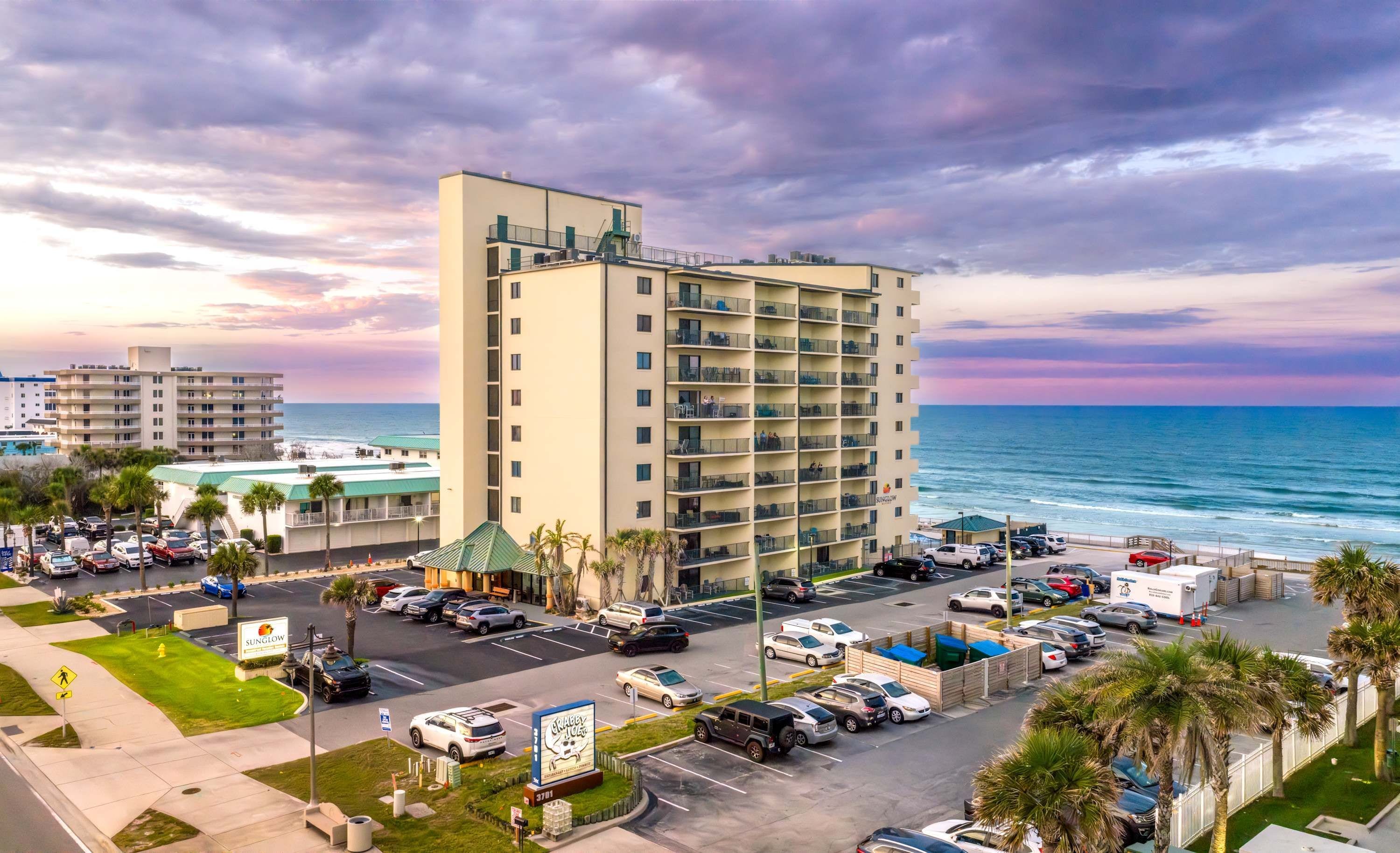 Um hotel moderno com várias varandas com vista para a praia, um estacionamento com diversos veículos e uma vista ampla do oceano.