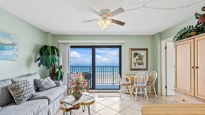 A living room with a sofa, a coffee table, a dining table with chairs, a ceiling fan, and a view of the ocean through large windows. - Sweet Sunrise Oceanfront Condo w Pool Hot Tub (Daytona Beach Shores)