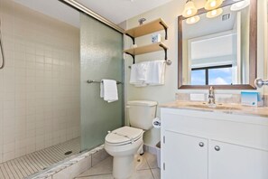 A bathroom with a shower, toilet, and vanity with a mirror and shelves. - Sweet Sunrise Oceanfront Condo w Pool Hot Tub (Daytona Beach Shores)