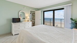 A bedroom with a bed, a dresser, a television, and a view of the beach through an open door. - Sweet Sunrise Oceanfront Condo w Pool Hot Tub (Daytona Beach Shores)