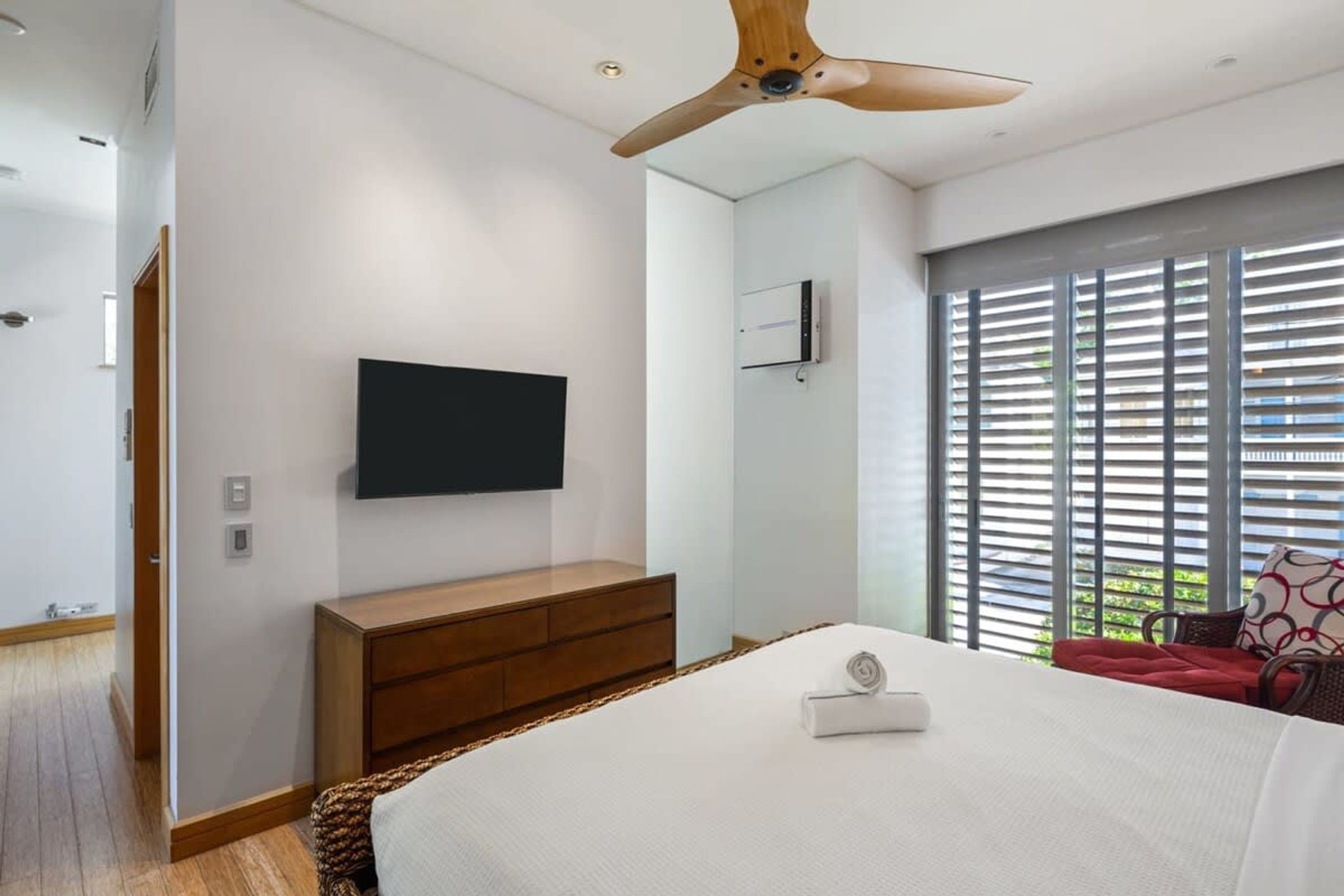 A modern bedroom with a bed, a ceiling fan, a TV mounted on the wall, and a window with blinds.