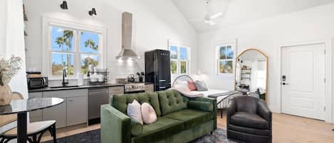 A spacious living area with a green sofa, a dark leather armchair, and a dining table with chairs. A kitchen with modern appliances and a large window is visible in the background.