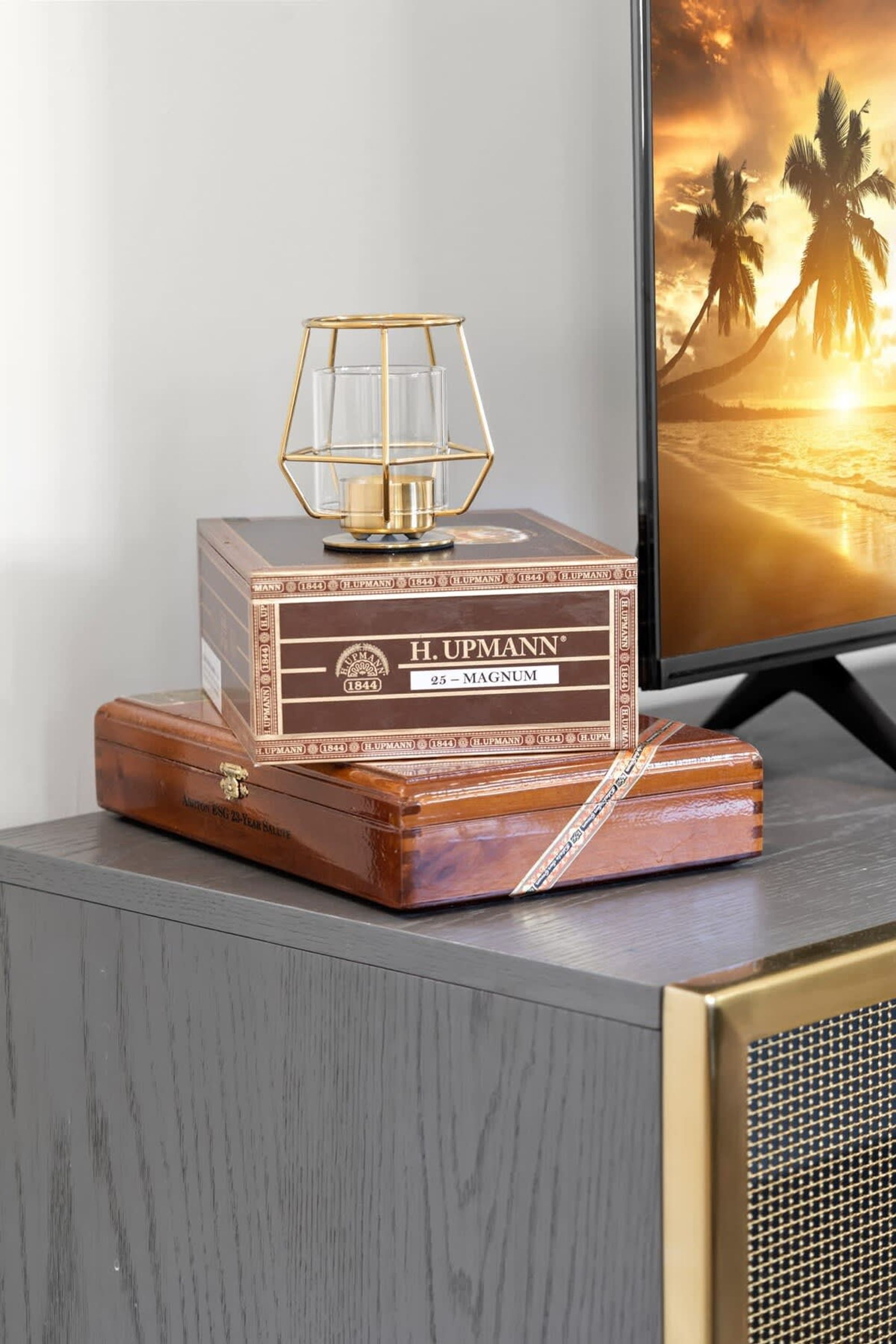 A wooden box with a glass lamp on top, labeled "H. UPMANN MAGNUM," placed on a grey surface next to a television displaying a sunset scene.