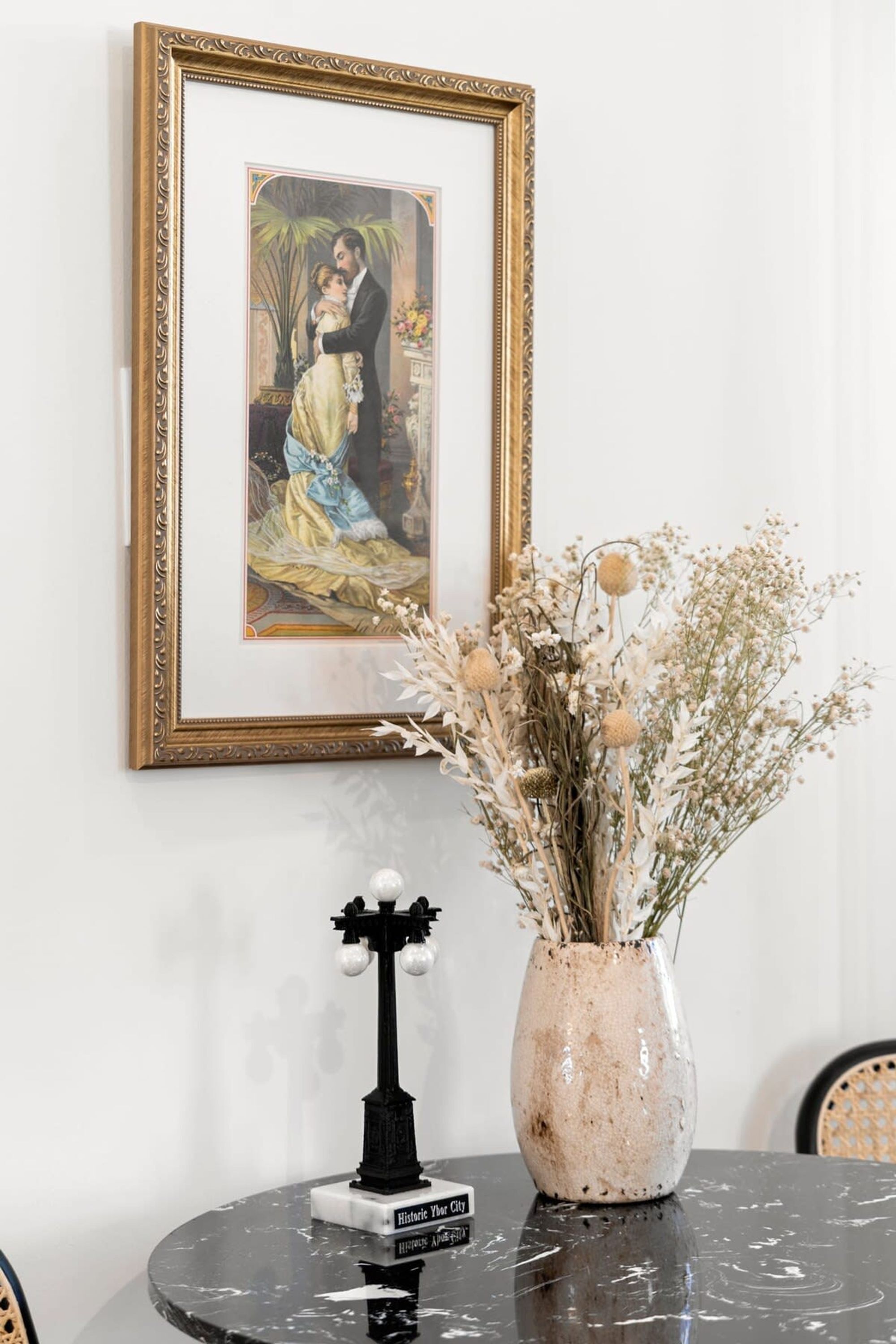 Ein gerahmtes Bild an der Wand, eine Vase mit Trockenblumen auf einem Tisch und ein kleines, schwarzes Objekt mit Etikett.