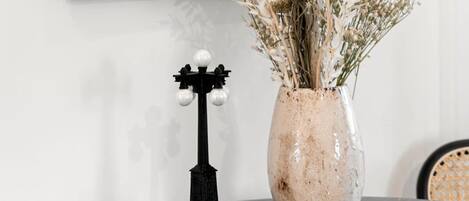 A framed painting on a wall, a vase with dried flowers on a table, and a small black object with a label.