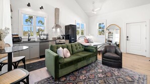 A spacious living area with a green sofa, a dark leather armchair, and a dining table with chairs. A kitchen with modern appliances and a large window is visible in the background. - Romeo Julieta 2 Epic Studios Steps to 7th Ave (Tampa)