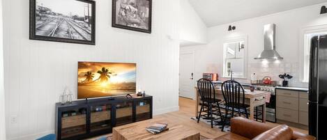 A modern living room with a wooden coffee table, a brown leather sofa, and a TV on a wooden cabinet. There is a dining area with a wooden table and chairs, and a kitchen with a black refrigerator and stainless steel range hood.