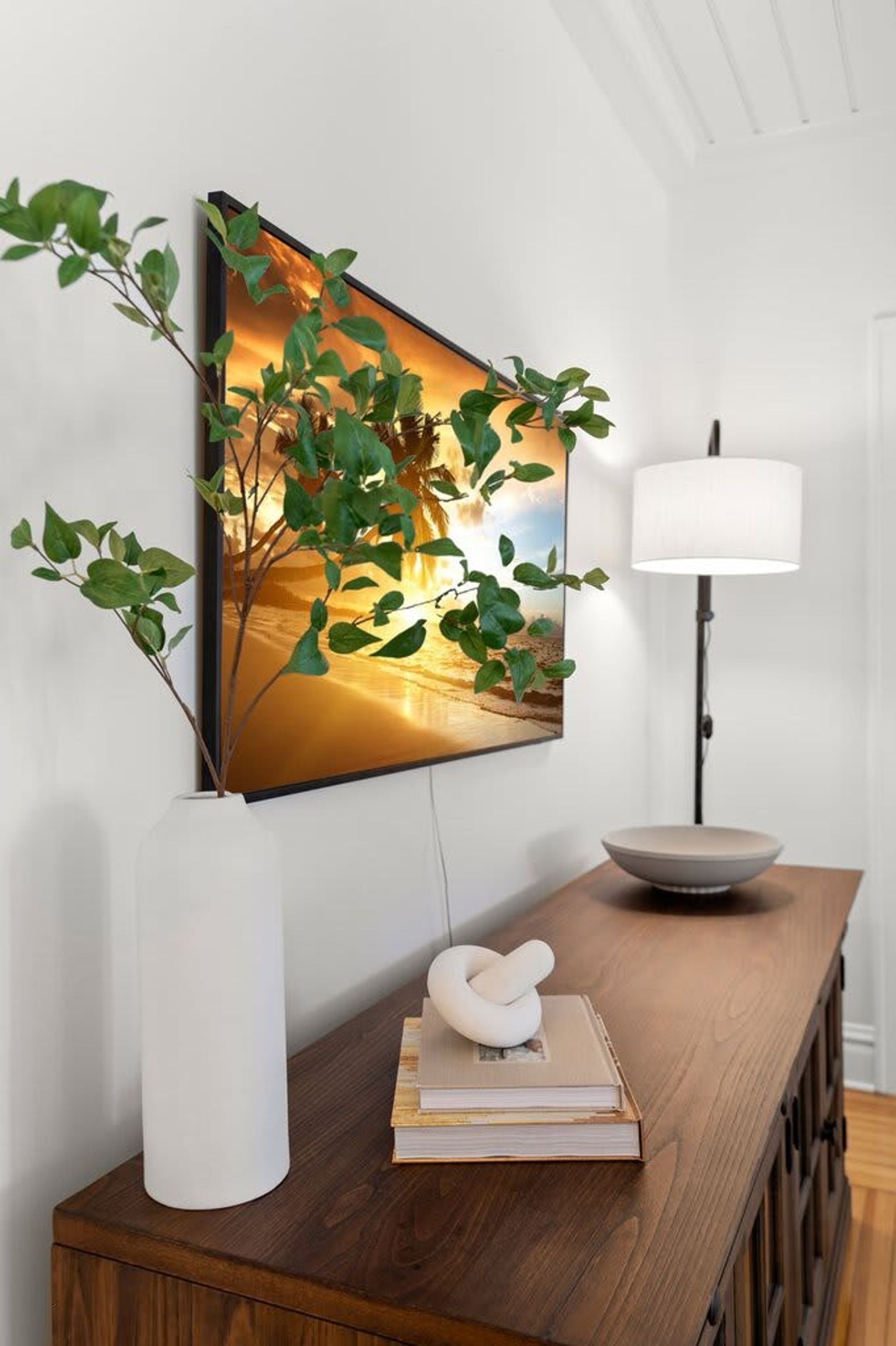 A modern living room with a wooden console table, a framed landscape painting, a white vase with green plants, a floor lamp, and a decorative sculpture.