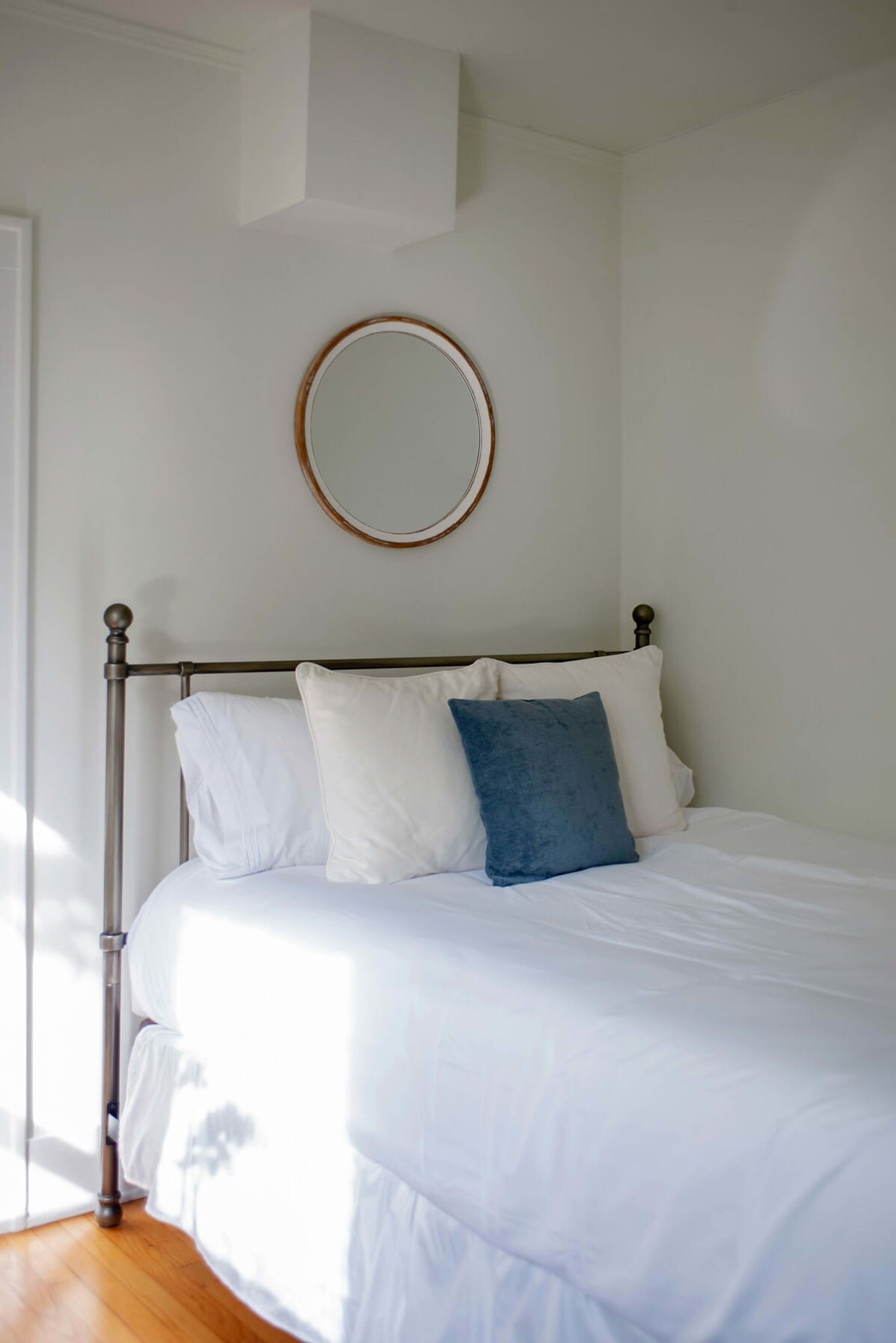 Una cama con ropa blanca y una almohada azul, un espejo redondo en la pared y suelo de madera.