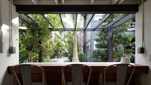 Ein Essbereich mit Holztisch und drei Stühlen, Blick auf einen Garten mit verschiedenen Pflanzen und einem großen Baum, sowie eine Pergola.