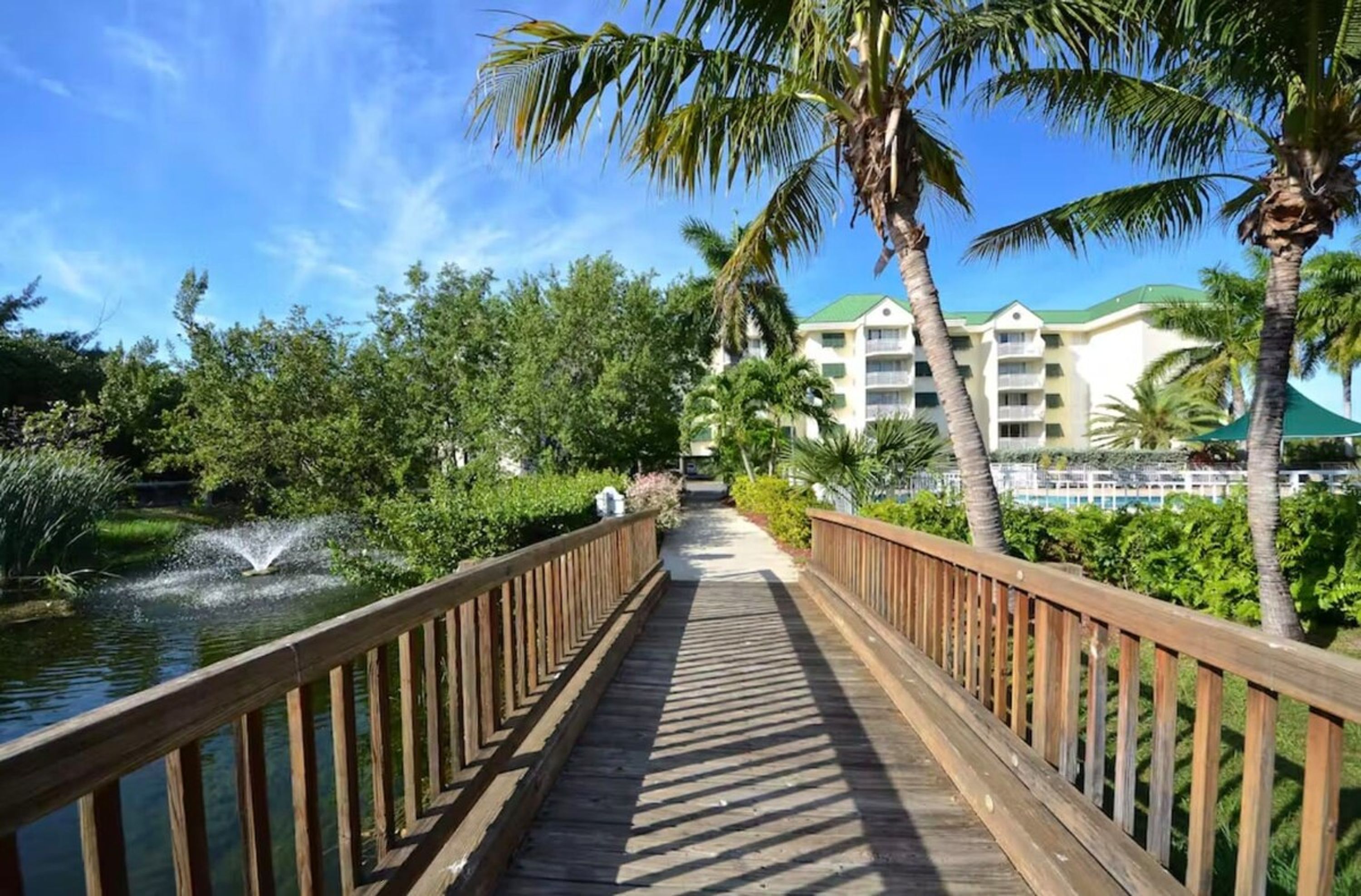 A wooden boardwalk leading to a building with a green roof, surrounded by palm trees and a body of water.